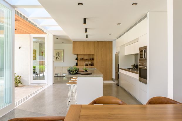 Bright open-plan kitchen and dining area with white built-in cabinetry, integrated stainless-steel appliances, a central island with bar stools, warm timber wall joinery, polished concrete flooring, and a timber dining table. Extensive glazing and a glazed courtyard connection provide abundant natural light.