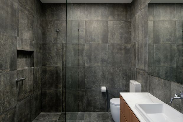 A compact, dark-toned contemporary bathroom with floor-to-ceiling large-format charcoal grey tiles covering the walls and floor. The room includes a frameless glass shower enclosure with recessed wall niche and exposed shower fittings, a white toilet, and a white integrated vanity basin with warm timber cabinetry. The bathroom has a moody, minimalist and materially cohesive appearance.