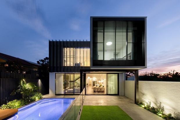 Twilight exterior view of a contemporary multi-level residence with strongly rectilinear forms, extensive floor-to-ceiling glazing, dark vertical facade fins, and a prominent projecting upper box with horizontal blinds. A lit dining area is visible through the ground-floor glazing. The property includes a swimming pool, glass balustrade, paved terrace, lawn, landscaped planting, and a rendered boundary wall.