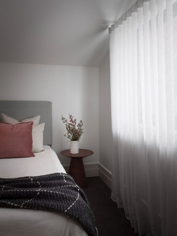 A softly lit bedroom corner with a partially visible bed, upholstered grey headboard, white bedding, and dusty-rose cushions. A dark charcoal knitted throw lies across the bed. A small dark timber pedestal side table holds a white vase with delicate branches, while full-height sheer white curtains filter daylight along the right wall. The room has white walls, layered white skirting boards, and dark carpet, creating a calm, understated atmosphere.