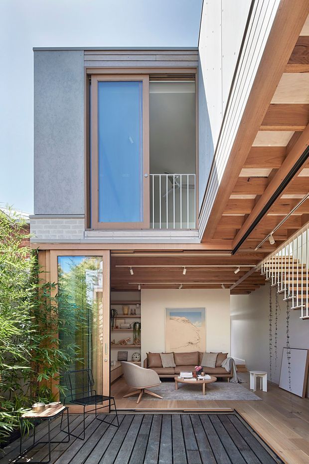 A bright, double-height courtyard living space viewed from a dark timber deck. The room features a warm timber-lined ceiling with exposed beams, large timber-framed glazing and doors, a contemporary sofa and lounge chairs, track lighting, and a white sculptural stair or balustrade structure. A small upper-level balcony with a frosted blue-glass door overlooks the living area. The composition blends indoor and outdoor spaces around a planted courtyard.