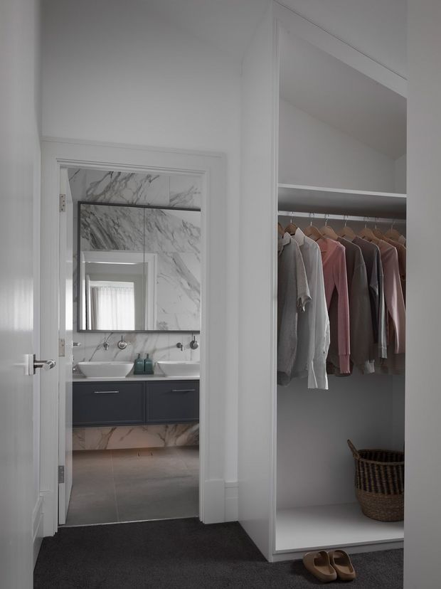 A refined dressing area with an open built-in wardrobe and hanging clothes, viewed toward an adjoining bathroom. The wardrobe has white joinery, a dark shelf and hanging rail, a woven basket, and shoes on a dark carpeted floor. Through the doorway, the bathroom features a white vessel basin, dark vanity cabinetry, large marble-look wall panels, a wide mirror, and grey floor tiles. The palette is predominantly white, grey, charcoal, and muted clothing tones.