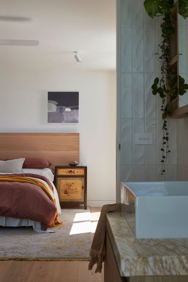 A warm bedroom viewed alongside an adjoining bathroom. The bedroom features a low timber headboard, layered mauve and mustard bedding, a patterned rug, and a vintage-style wooden bedside cabinet. The bathroom has pale blue-green glazed wall tiles, a freestanding or built-in bathtub edge in the foreground, a stone-patterned vanity surface, and trailing plants on open shelving. Soft daylight and minimal white walls create a relaxed, eclectic atmosphere.