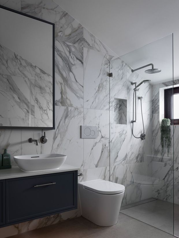 A sophisticated contemporary bathroom featuring dramatic white marble-effect wall tiles with grey veining, a dark navy or charcoal floating vanity with a white vessel basin, wall-mounted tapware, a concealed cistern toilet, and a frameless glass walk-in shower with dual showerheads. A large black-framed mirror and small window with trailing greenery complete the refined, high-contrast scheme.