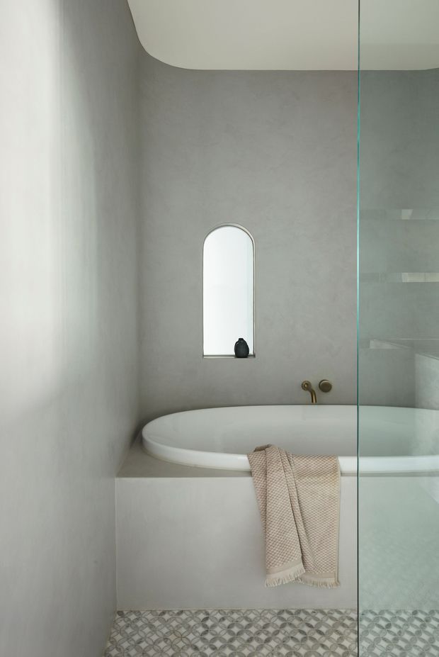 Minimalist bathroom featuring a built-in oval bathtub, soft grey plastered walls, a narrow arched recessed mirror or window, brushed brass wall-mounted tapware, a frameless glass shower screen, and patterned geometric floor tiles. The space has a calm, refined, monochromatic appearance with a subtle warm textile accent.