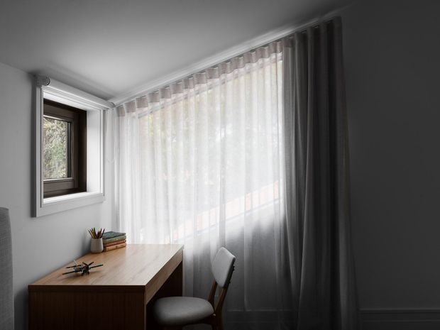A compact, softly lit home office with a built-in or freestanding timber desk positioned beside a large window. Sheer white curtains and dark grey drapes filter daylight, while a simple upholstered chair, small books, and stationery create a restrained workspace. The room has white walls, a plain ceiling, and classic skirting and window trim.