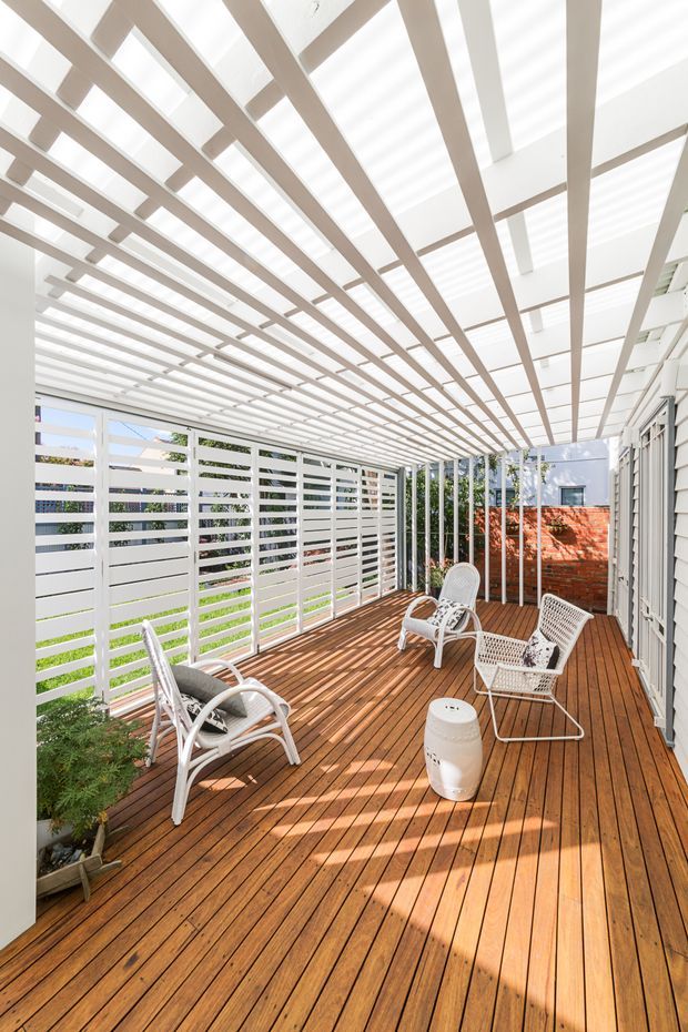 A bright covered outdoor terrace or deck with warm timber decking, white horizontal privacy screens, slender white posts, and a distinctive overhead pergola with translucent roofing or skylight panels. White outdoor lounge chairs and a small side table are arranged along the deck, with greenery and a brick boundary wall visible beyond.
