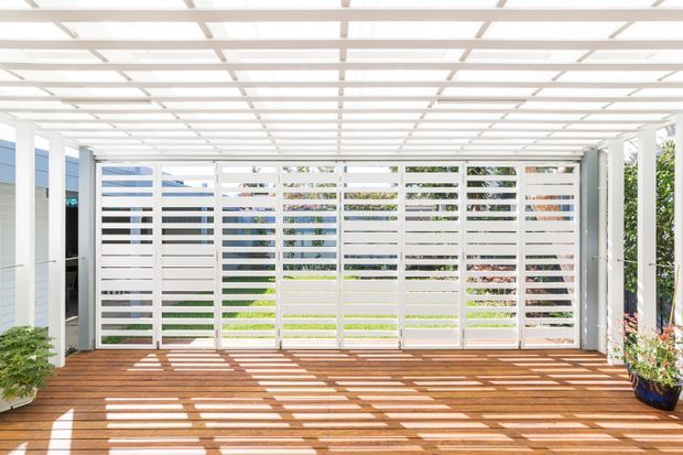 Bright enclosed outdoor room or covered deck with warm timber decking, white horizontal slatted screening, and a translucent or open-grid overhead pergola roof. Strong linear sunlight and shadows fall across the deck, with garden greenery visible through the screens and potted plants at the edges.