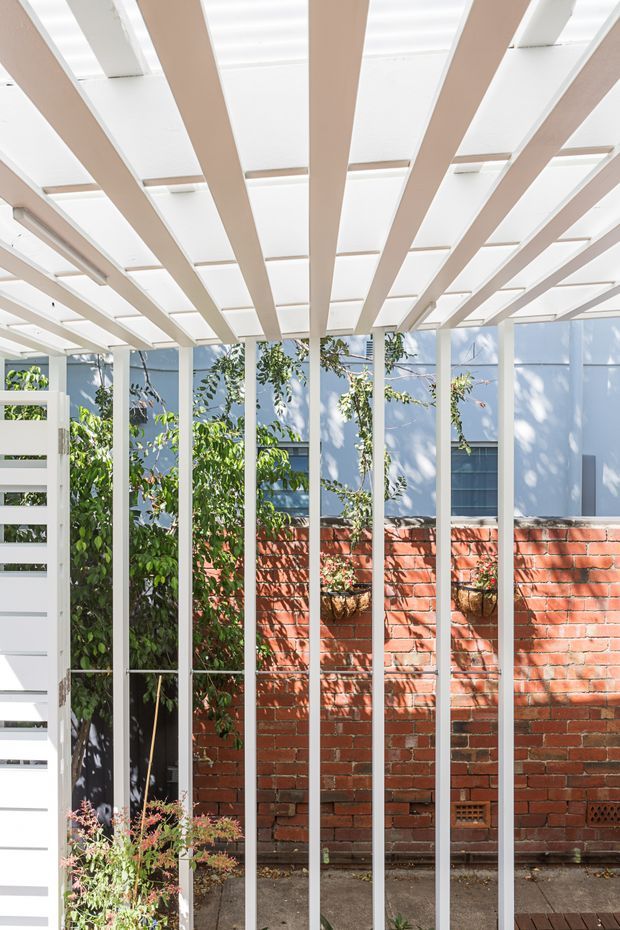 Bright architectural view of a compact courtyard or covered outdoor walkway, framed by a white pergola with regularly spaced beams and translucent roof panels. Slim white vertical screens create a rhythmic enclosure, with a white slatted gate at left. A red brick boundary wall, blue exterior wall, climbing greenery, and hanging planters are visible beyond. The composition is airy and geometric, with strong daylight and patterned shadows.