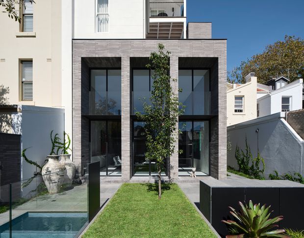 Rear facade of a contemporary multi-storey residence viewed from a landscaped courtyard. The building combines pale rendered walls with a dark grey brick-clad projecting volume containing large black-framed floor-to-ceiling windows and glazed doors. A narrow lawn, glass-edged reflecting pool, paved paths, raised planter, and sculptural planting define the foreground courtyard.