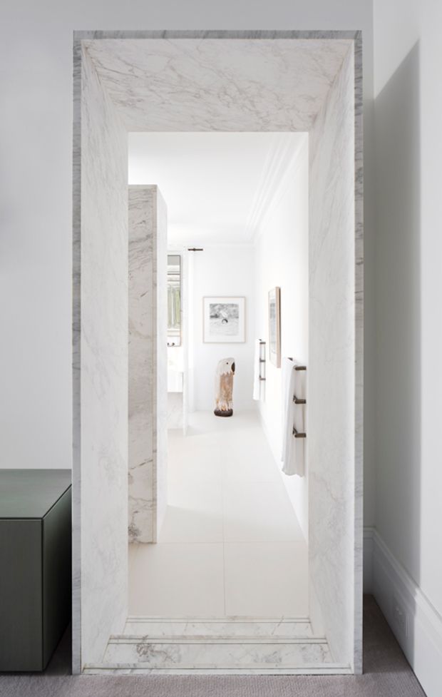 A bright, minimalist interior view through a substantial white-veined marble-clad doorway into a narrow white hallway or bathroom-like space. The opening has marble-lined reveals and threshold detailing, white tiled flooring, white walls and ceiling, a wall-mounted towel rail on the right, framed artwork, and a sculptural object at the far end. A dark built-in cabinet is partially visible outside the opening on the left, with grey carpet and white skirting in the foreground.