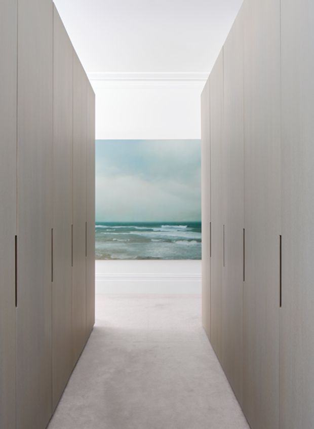 A narrow, symmetrical hallway lined on both sides with tall, handleless light-toned built-in cabinetry or wardrobes. The corridor leads toward a large ocean artwork or seascape image framed by white walls and decorative cornicing. The space has a soft, monochromatic palette, pale carpet or smooth flooring, and a bright, serene presentation.