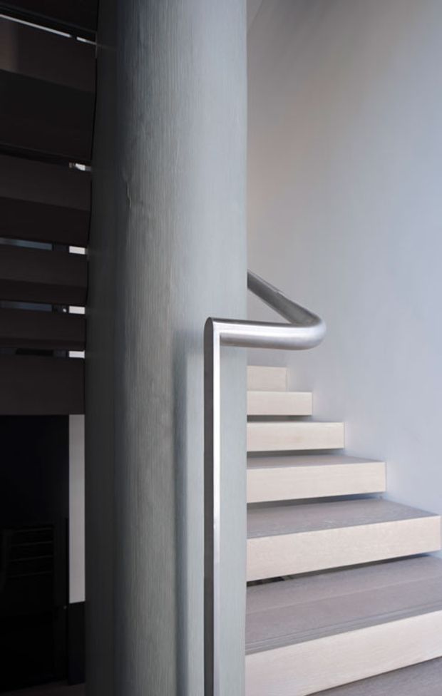 Close-up interior view of a contemporary stairway with pale stone or concrete treads, a smooth light wall, a prominent cylindrical structural column, and a curved brushed-metal handrail. Dark horizontal screening or slatted architectural elements appear to the left, creating a strong contrast with the bright stairwell.
