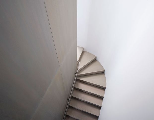 A minimalist spiral stairway viewed from above, with tapered light timber or wood-look steps wrapping around a central wall or structural core. The stair is enclosed by smooth white walls, with a slim dark metal handrail and restrained detailing. The composition is clean, sculptural, and softly lit.
