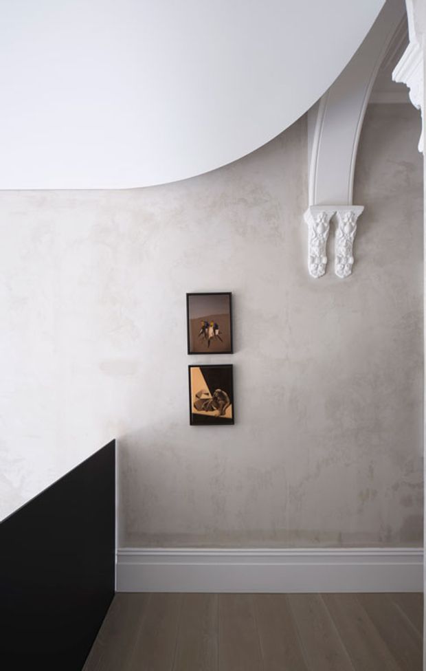 A refined interior corner featuring a curved white ceiling transition, pale textured plaster walls, ornate white classical arch detailing with decorative corbels, white panelled skirting, and a dark timber floor. A black geometric stair or partition element occupies the lower left, while two small framed artworks hang vertically on the wall.