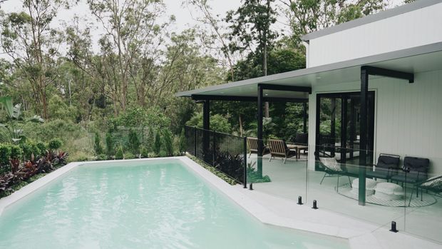 Contemporary modernist home beside a pale turquoise swimming pool, set within a lush wooded landscape. The building features crisp white horizontal cladding, extensive black-framed glazing, a flat roof with broad overhangs, and a covered outdoor terrace furnished with lounge chairs. A black metal pool fence and light-coloured paved pool surround define the outdoor area.