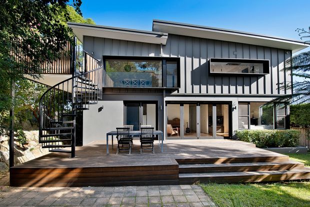 A contemporary two-storey home viewed from the rear garden, with grey vertical cladding, expansive glazed doors and windows, and a broad timber deck with stepped stairs. A distinctive black spiral staircase connects the deck to the upper level. The landscaped garden and paved path are visible in the foreground.