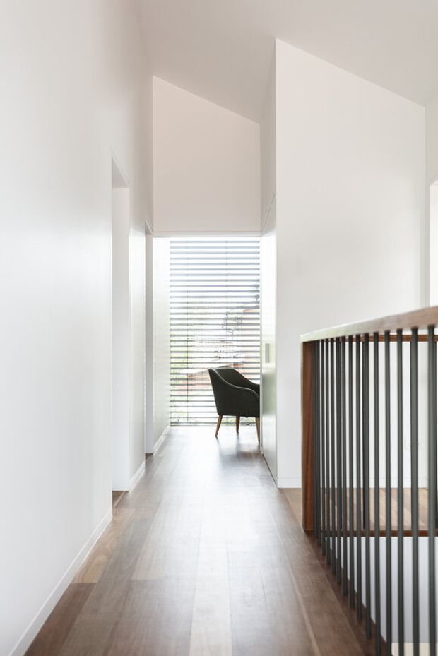 Bright minimalist interior hallway or landing with pale walls, timber flooring, and a dark vertical-slat stair balustrade with timber handrail. A lounge chair sits at the end near a large window fitted with horizontal blinds, creating a calm, light-filled circulation space.