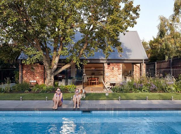 A contemporary residential backyard with a long rectangular swimming pool in the foreground and a compact gable-roofed house behind it. The house combines exposed red brick walls, dark corrugated metal roofing and a broad glazed opening to a timber-decked outdoor living area. Mature trees, planted garden beds, a lawn strip and dark boundary fencing surround the pool and house.