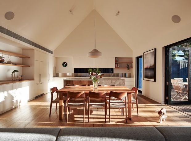 Warm, open-plan dining and kitchen interior with a large timber dining table and matching chairs in the foreground. The kitchen has white full-height cabinetry, open timber shelving, a dark horizontal splashback, and a stone-look island or benchtop. A vaulted ceiling with a central pendant light adds height, while large glazed doors open to an outdoor deck. Timber flooring, abundant natural light, framed artwork, and a small dog complete the relaxed residential setting.