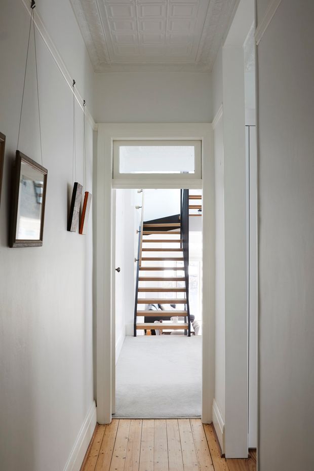 A narrow, bright residential hallway with a warm timber floor, white walls, decorative ceiling moulding, framed artwork and a doorway leading to an open-riser staircase. The space has traditional detailing combined with a clean, contemporary white palette.