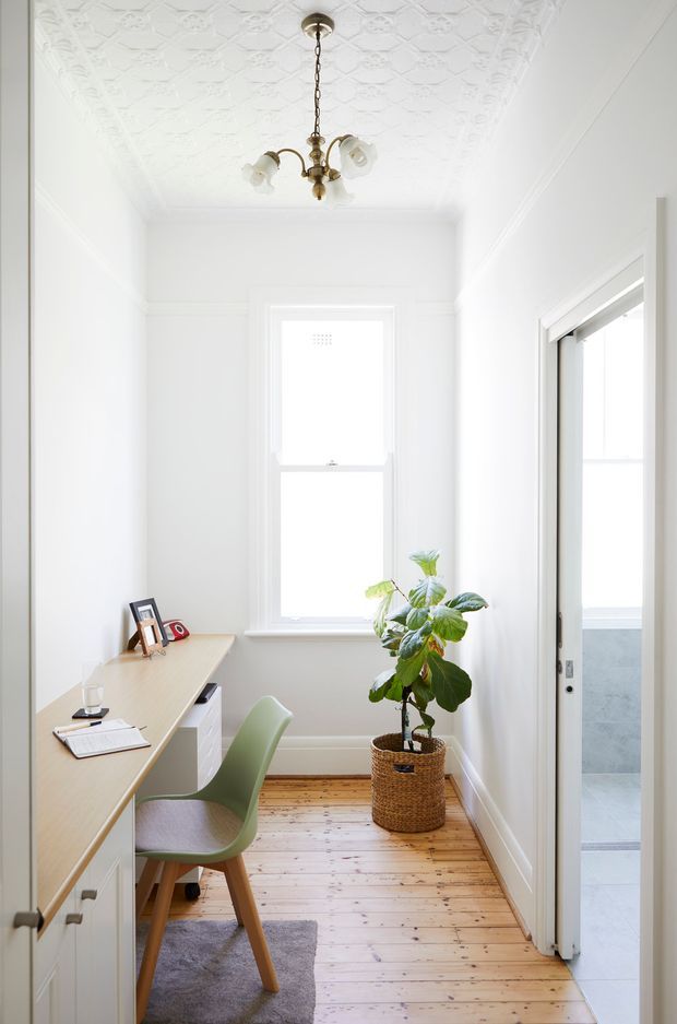 Bright, narrow home office with white walls, a decorative pressed-tin patterned ceiling, timber floorboards, and a compact built-in desk with drawers along the left wall. A pale green chair sits at the desk, with framed photos and stationery on the work surface. A potted plant in a woven basket occupies the window area, and an open doorway reveals a tiled adjacent room. The room has a traditional character with abundant natural light and a vintage brass pendant light.