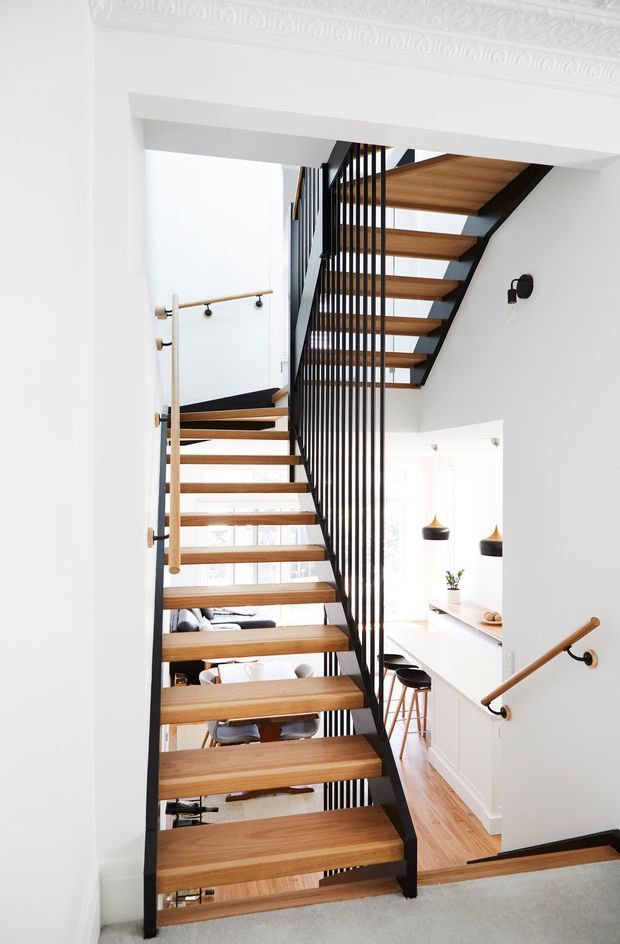 Bright contemporary stairway with open-riser timber treads, black steel stringers and vertical balustrade, and timber handrails. The stair connects multiple levels and overlooks a light-filled kitchen and dining area. White walls feature ornate decorative cornice moulding, creating a refined contrast with the modern stair construction.