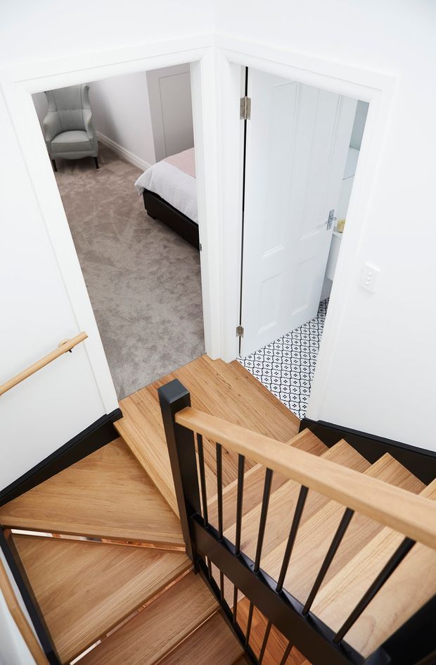 Bright, white-painted stairwell viewed from above, with natural timber stair treads and landing, black metal balusters and stringer, and a timber handrail. Open doors reveal a carpeted bedroom with a bed and armchair on the left and a tiled bathroom on the right. The stair geometry and contrasting timber-and-black railing dominate the image.