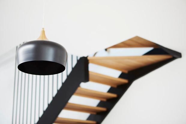 Close-up interior view of a contemporary open-riser staircase with warm timber treads, a dark structural stringer, and slim vertical metal balustrade rods. A large black and brass pendant light hangs in the foreground against a clean white wall.
