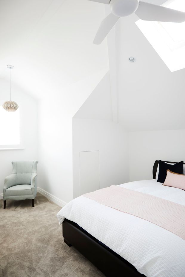 Bright, predominantly white bedroom with a sloped ceiling and skylight, featuring a ceiling fan, plush beige carpet, a black upholstered bed with white and blush bedding, and a pale upholstered armchair beside a window. A geometric pendant light and concealed wall storage add subtle detail.
