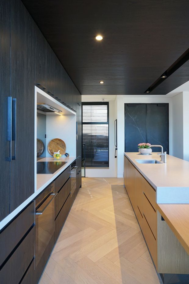A narrow, contemporary galley kitchen with dark wood full-height cabinetry, integrated appliances and open shelving along one wall, and a large light-toned island with an undermount sink and modern tap. The room features a dark timber ceiling with recessed and linear lighting, pale herringbone timber flooring, a black-framed window with horizontal blinds, and a dark internal door. The palette is sophisticated and strongly contrasted, combining dark timber, light wood, white surfaces, and black accents.
