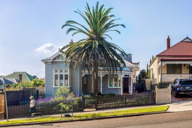 Street view of a compact heritage-style timber cottage with a pale blue-grey facade, ornate white fretwork, decorative window trims, a front veranda, and a steeply pitched roof. A mature palm tree dominates the fenced front garden, with neighbouring period homes and a driveway visible to either side.
