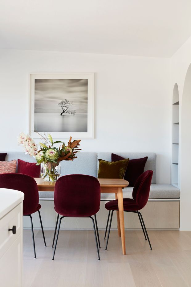 A bright, refined dining area with a light timber dining table, built-in pale upholstered bench seating, and deep burgundy velvet dining chairs. The room has white walls, pale timber flooring, recessed shelving, a large monochrome landscape artwork, and a floral arrangement as a focal point.