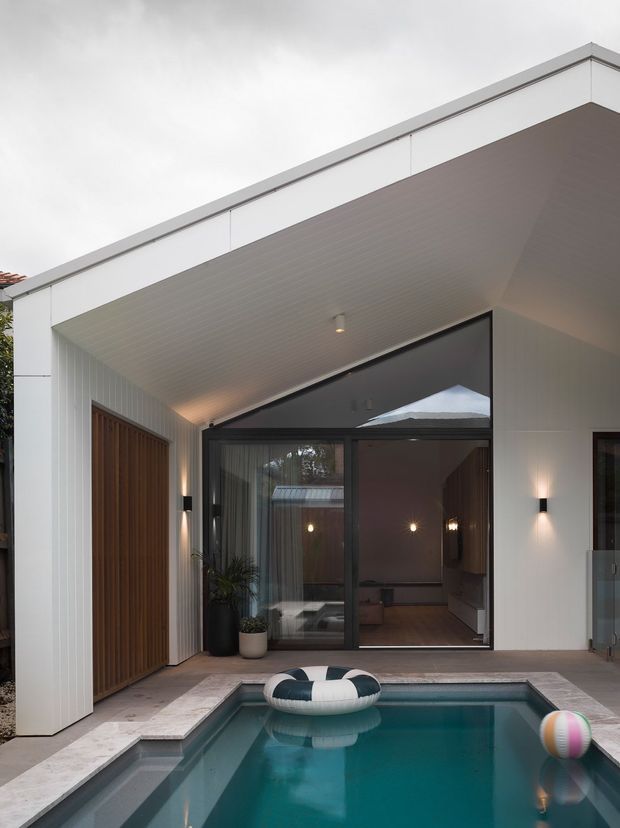 A contemporary residential courtyard viewed from beside a compact swimming pool. The home has a crisp flat-roofed white facade, deep covered outdoor opening, vertical wall cladding, dark-framed full-height glazing, and a tall timber front door. Warm wall sconces and interior lighting create contrast against the cool, overcast exterior. The pool occupies the foreground with floating inflatables, while a living area is visible through the glass doors.
