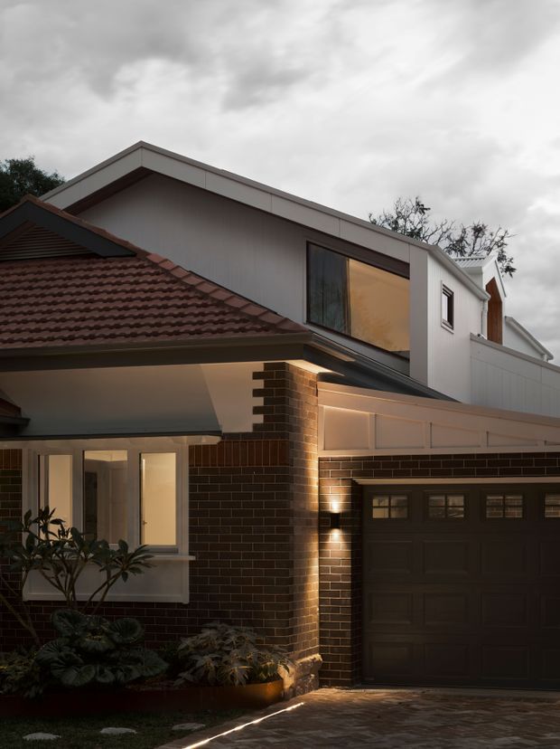 Exterior photograph of a two-storey suburban home at dusk, showing a modern upper-level extension above an older tiled-roof brick dwelling. The facade combines dark brown brickwork, light rendered wall panels, white trim, a large upper window, a covered entry, and an integrated dark garage door. Warm wall lighting illuminates the brick column and paved driveway, with planting along the front edge.