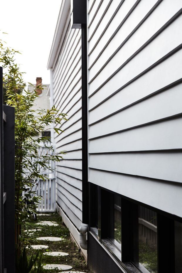 A narrow side passage between residential buildings, featuring a white horizontal weatherboard-clad wall with black window frames and downpipes. Circular stepping stones lead through a small landscaped strip with bamboo-like planting toward a rear gate. The scene has a crisp, contemporary residential exterior character with strong linear perspective and high contrast between white cladding and dark trim.