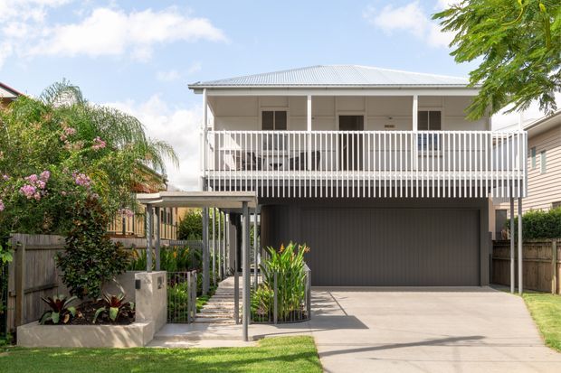 Front exterior view of a raised two-storey residential home with a light weatherboard facade, broad upper-level balcony with white vertical balustrades, a corrugated metal roof, and a dark enclosed garage beneath. A concrete driveway, covered pedestrian entry, fencing, and landscaped planting frame the facade.