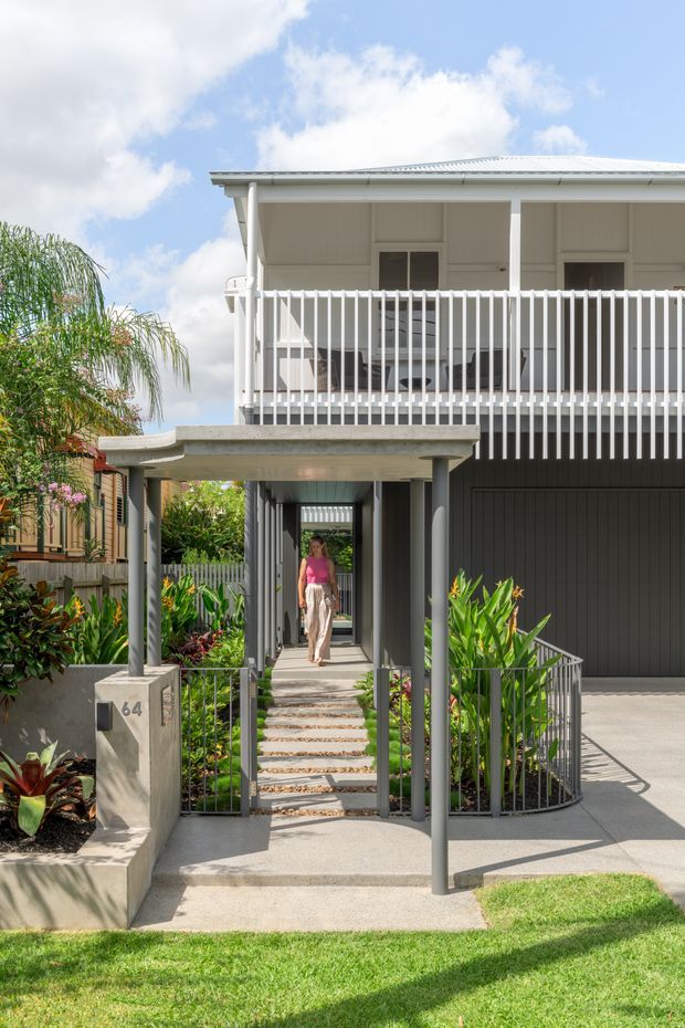 A bright street-facing view of a raised two-storey residential home with a deep covered entry walkway, upper-level balcony, vertical white balustrades, pale weatherboard-style walls, and a light metal roof. The lower level includes a dark garage door, while landscaped tropical planting, steps, and a front lawn frame the entrance.