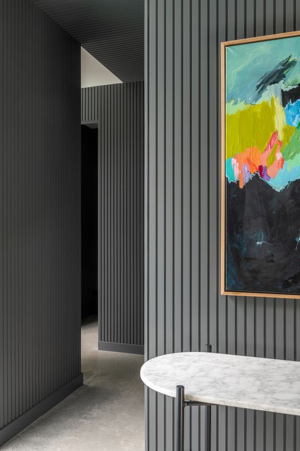 A sophisticated modern interior corridor or entry space featuring dark vertical fluted wall panelling that continues across the ceiling, a polished concrete floor, and a round white marble table with a slim dark metal base in the foreground. A colourful abstract artwork in a timber frame provides a vivid accent against the charcoal walls. The space is moody, refined, and minimalist with strong linear detailing.