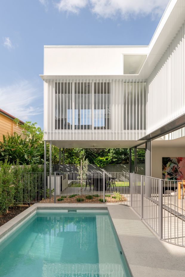 Exterior view of a bright contemporary multi-level home centered around a swimming pool. The white upper storey is elevated on slender supports and enclosed by dense vertical screening, with large glazed openings and a flat roof. A covered outdoor area, metal balustrades, landscaped planting and pool surround are visible at ground level.
