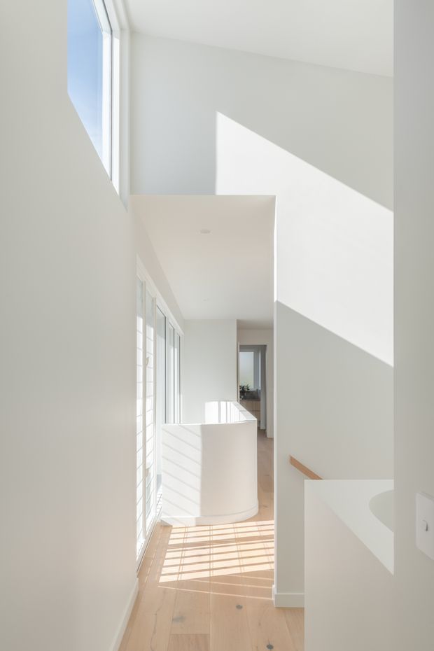 A bright, minimalist interior circulation space viewed from a stair landing or upper hallway. White walls and a white balustrade frame a light timber floor, while tall glazing casts strong geometric sunlight and shadows across the floor and walls. A stair opening and timber handrail are visible on the right, with a distant doorway and glimpse of greenery beyond.