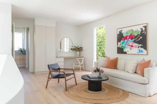 Bright, calm living room with a pale timber floor, white walls and built-in cabinetry. The room includes a large white sofa with warm terracotta cushions, a low black round coffee table, a woven circular rug, a timber-framed lounge chair, and a compact writing desk beneath an arched mirror. A tall louvred window brings in greenery and daylight, while an open doorway leads toward another interior area.
