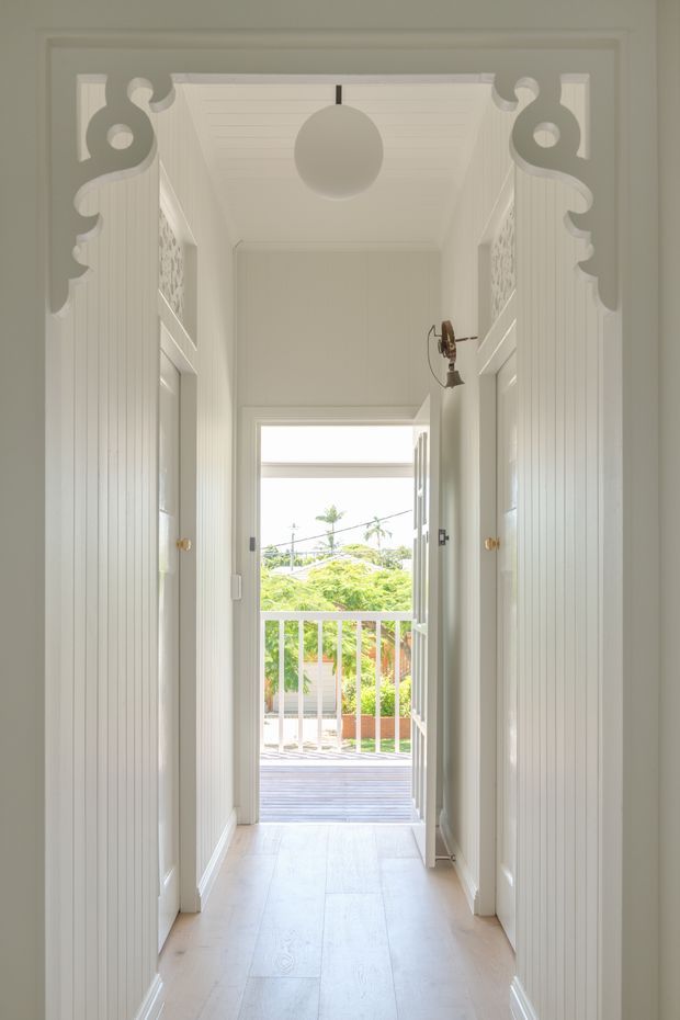 A bright, narrow residential hallway lined with white vertical-panel built-in cupboards and decorative carved arch details at the upper corners. The corridor has pale timber flooring, a white paneled ceiling, a central globe pendant light, and a small wall light. Open double doors at the end lead to a timber deck or balcony with a white balustrade and leafy garden beyond.