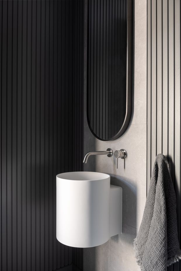 Close-up of a contemporary bathroom vanity featuring a cylindrical white wall-mounted basin, brushed metal wall-mounted tapware, a tall rounded-edge black mirror, and dark vertical ribbed wall panelling contrasted with light stone-look wall surfaces. A textured grey towel hangs on the right.