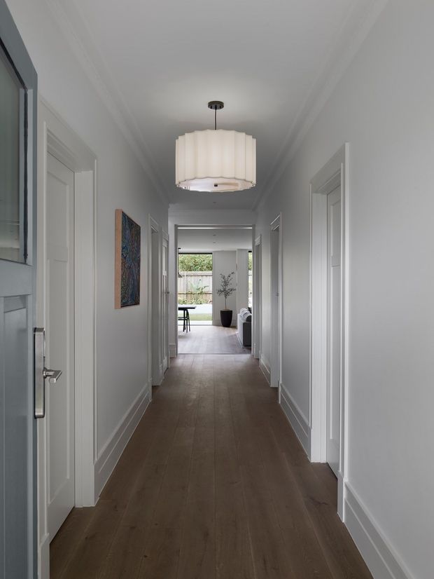 A long, bright residential hallway with white paneled doors, crisp white walls and trim, dark timber-look flooring, and a central pleated pendant light. The corridor leads toward an open living area and garden-facing glazing at the far end, with a framed artwork and a small plant adding limited colour.