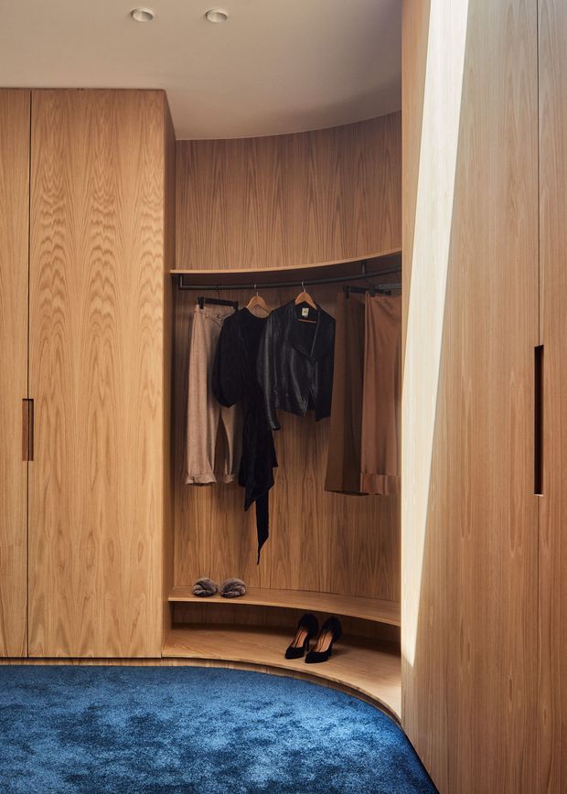 A compact, highly finished walk-in wardrobe with curved built-in timber joinery, hanging rails, shelves, and concealed storage. Warm wood grain cabinetry contrasts with a deep blue carpet, while recessed ceiling lights provide soft illumination. Clothing and shoes are visible within the open wardrobe section.