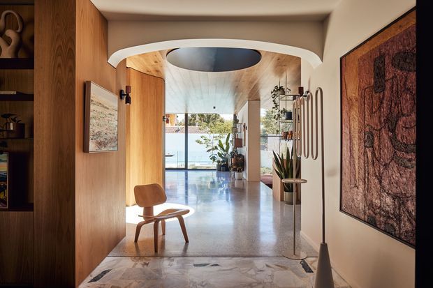 A refined residential entry hall opens toward a bright glazed living area. The space features warm timber wall panelling and built-in shelving, a sculptural curved plaster ceiling transition with a circular recessed opening, polished light stone or concrete flooring, and a large abstract wall artwork. A timber lounge chair, plants, decorative objects, and a sculptural floor lamp add mid-century-inspired character. Full-height glazing at the far end brings in abundant natural light and views of greenery and water or sky.