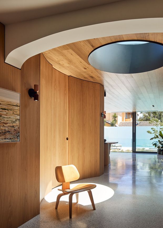 A refined residential interior entry or circulation space featuring curved oak-toned timber wall panelling and a sculptural curved ceiling with a large circular skylight or void. A warm wooden lounge chair sits in a pool of sunlight on a polished terrazzo-like floor. Full-height glazing opens views toward a bright outdoor pool or courtyard, while integrated wall lights and minimal furnishings reinforce the carefully detailed contemporary design.