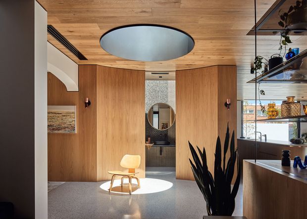 A refined contemporary interior with warm timber lining across the ceiling and full-height wall panels, opening toward a compact bathroom or powder room. A sculptural circular ceiling light, curved white architectural detail, terrazzo-like flooring, and built-in timber joinery create a polished, minimalist composition. Open shelving and a timber kitchen or bar counter are visible on the right, with indoor plants and a small sculptural lounge chair adding softness.