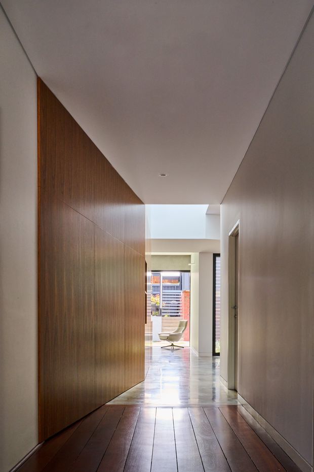A long, narrow residential hallway with a warm dark timber-panelled built-in wall on the left, smooth light-painted walls and ceiling, recessed downlighting, and dark timber floorboards in the foreground. The corridor opens toward a bright living area with polished light flooring, a lounge chair, and visible exterior glazing or courtyard beyond. The composition emphasises the strong linear perspective, contrasting timber surfaces, and a high ceiling transition near the end of the hall.